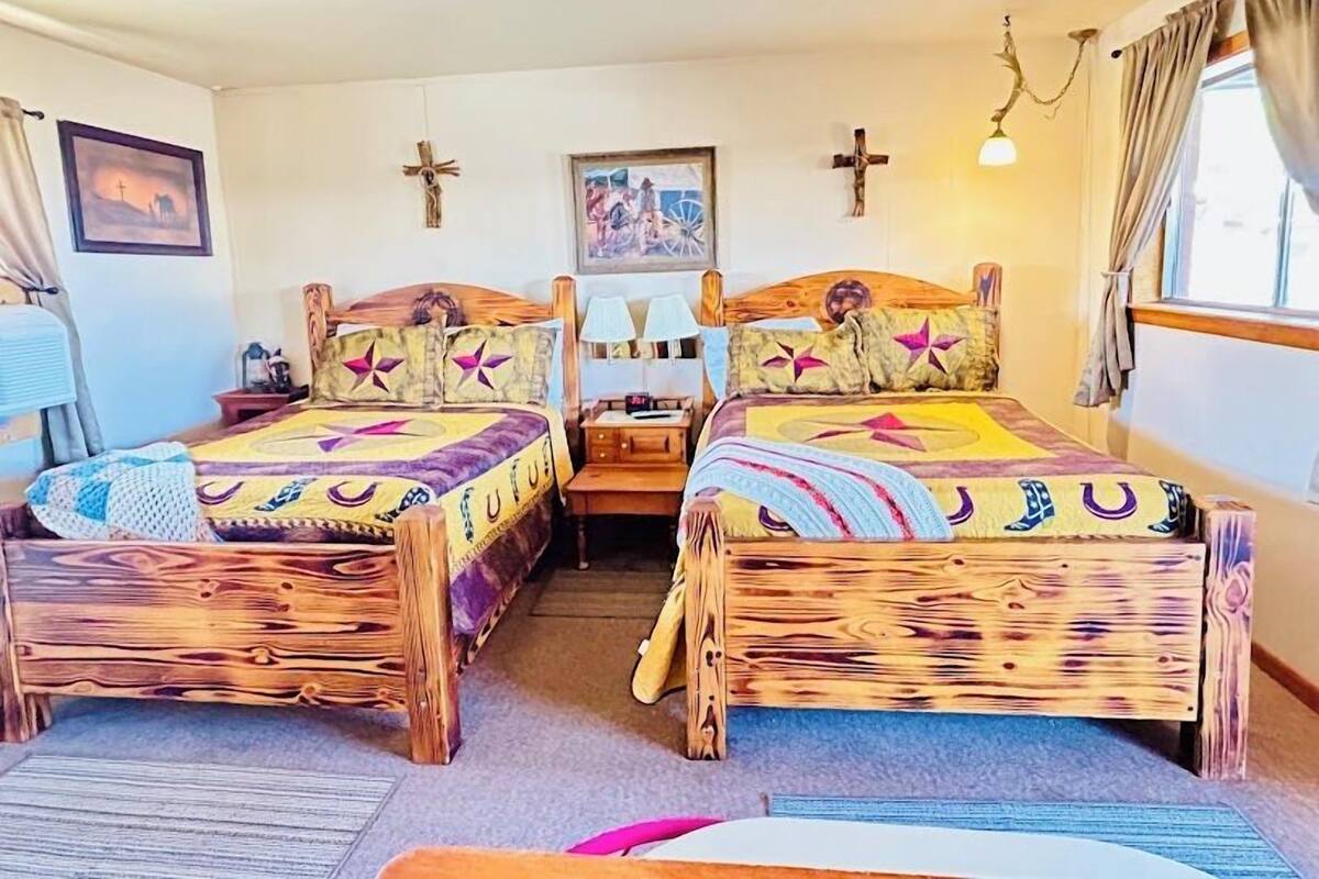 A room with two wooden beds, each with a unique quilt design. There are wall-mounted crosses and a framed picture above the beds.