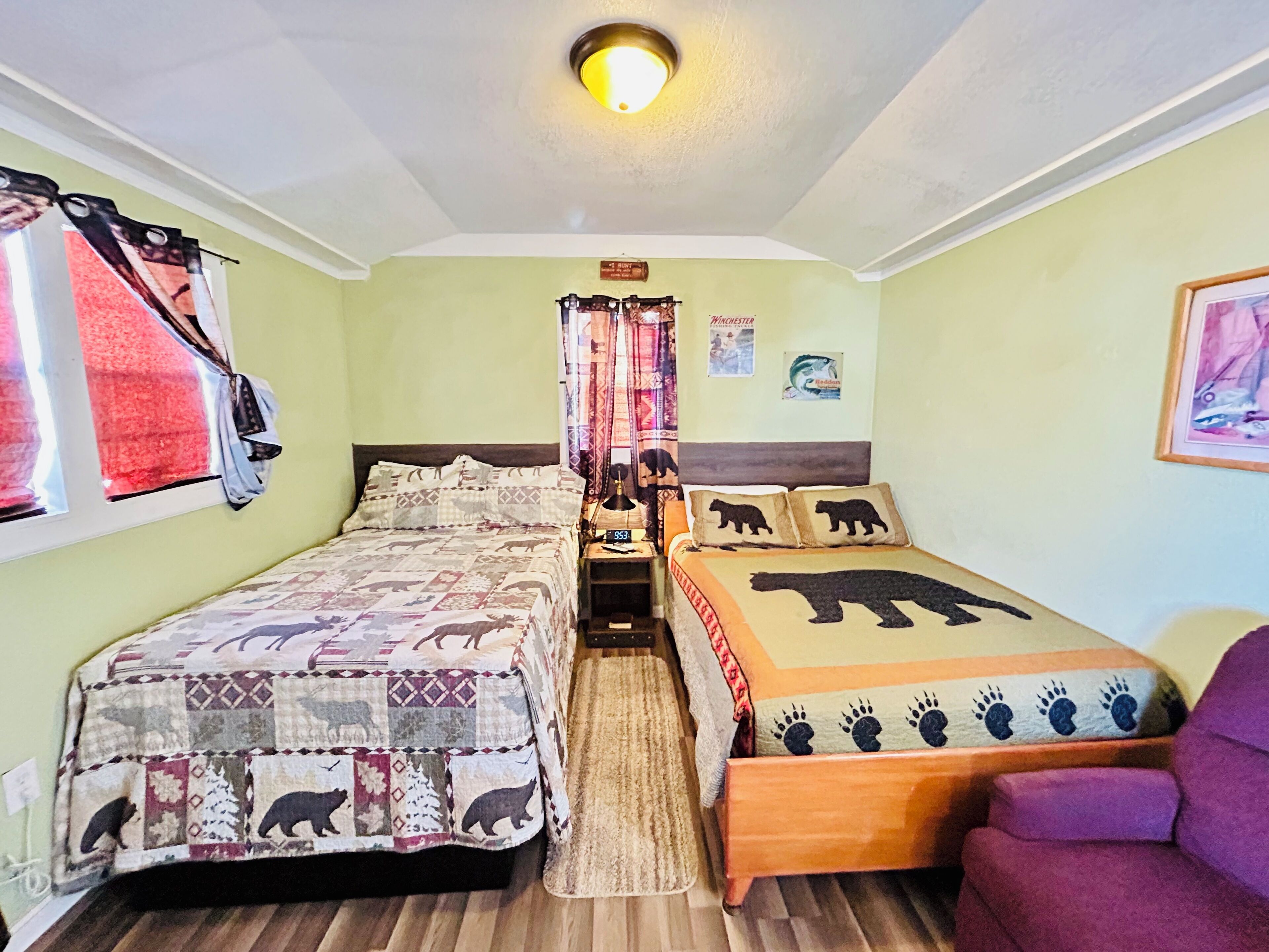 Una habitación con dos camas, una con diseño de oso acolchado y otra con huellas de pata. Se ve una silla morada y una ventana con cortinas a la izquierda.
