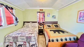 Una habitación con dos camas, una con diseño de oso acolchado y otra con huellas de pata. Se ve una silla morada y una ventana con cortinas a la izquierda.