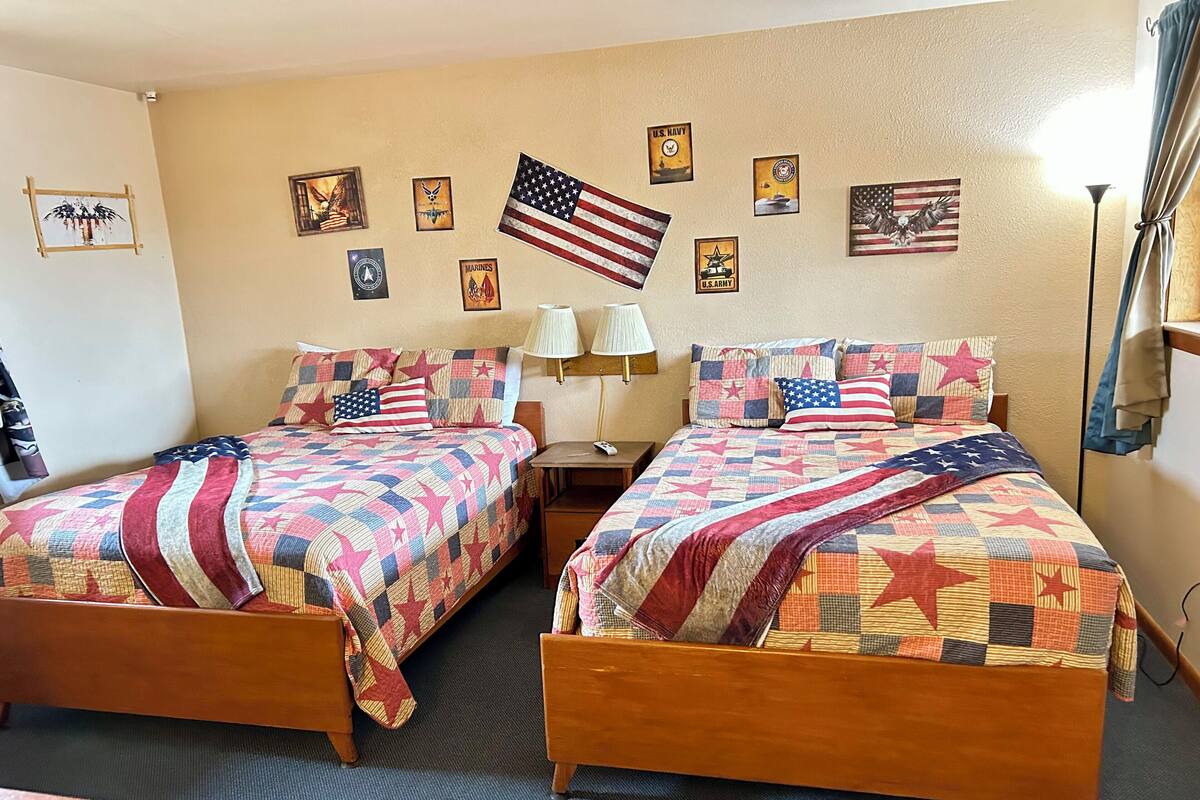 A hotel room with two beds, each adorned with a quilt featuring a star pattern. There are multiple framed military insignia on the wall, including the U.S. Navy and Marines.
