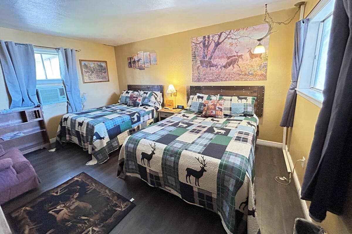 A room with two beds, a wooden dresser, a rug, and a painting of deer on the wall.