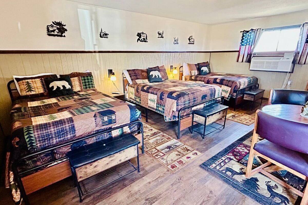 A room with two beds, each with a quilted blanket and pillows, a small table, and a chair.