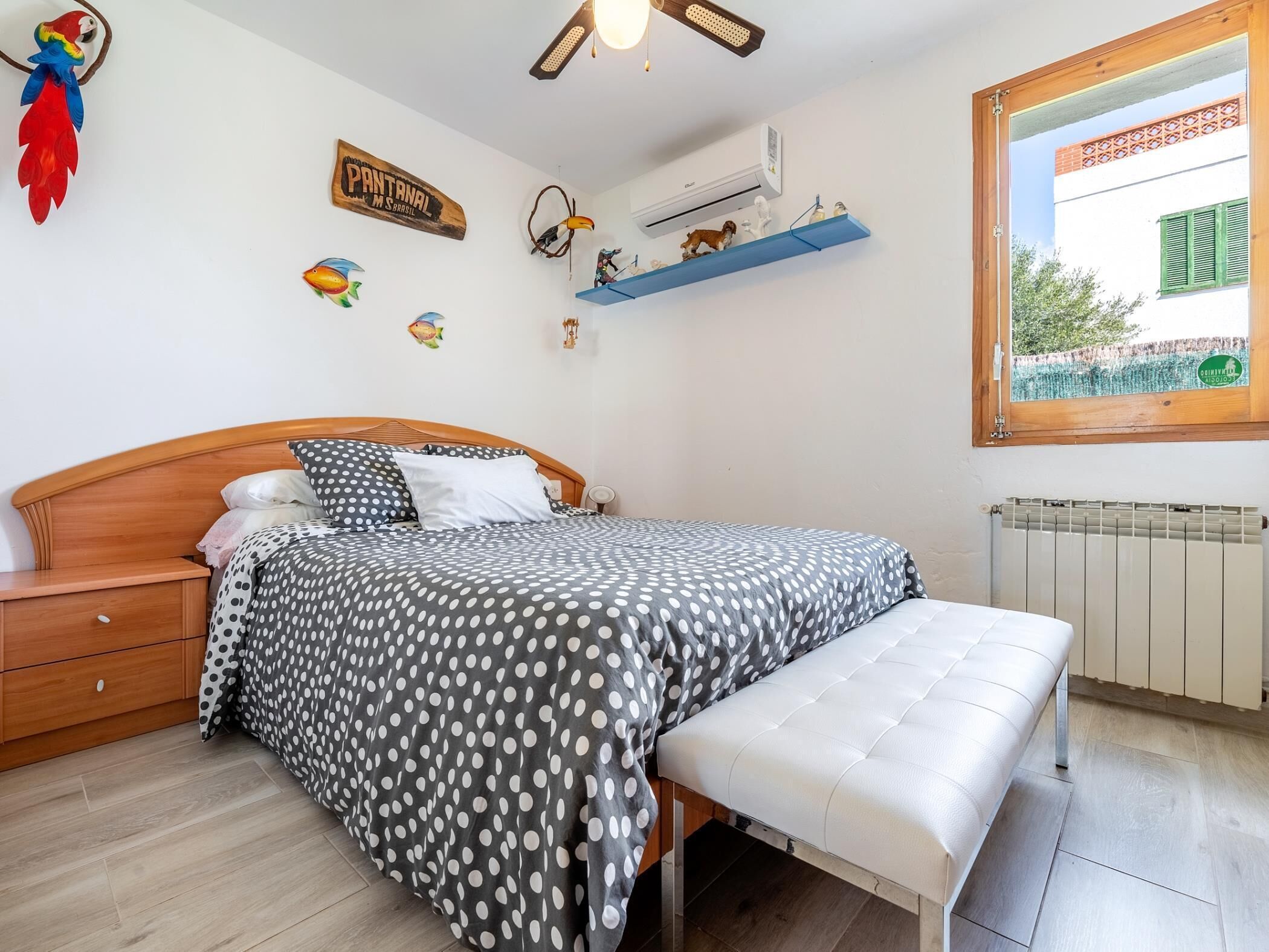 Una camera da letto con un letto, una panca, un comodino, una mensola a parete con figurini, una sveglia a muro e una finestra vista sul verde.