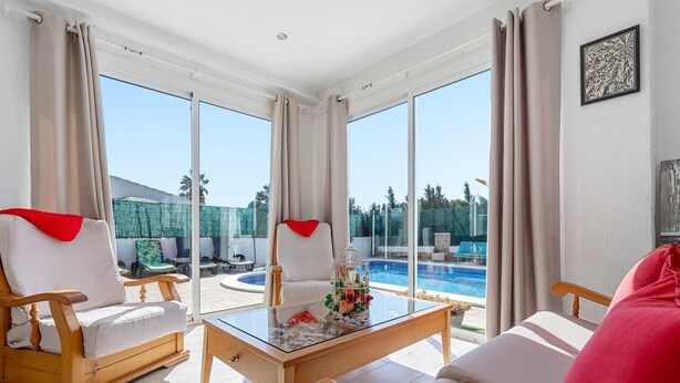 Amplia sala de estar con una mesa de vidrio, dos sillones y un sofá. Un gran ventanal ofrece vistas a una piscina y un área de estar exterior.