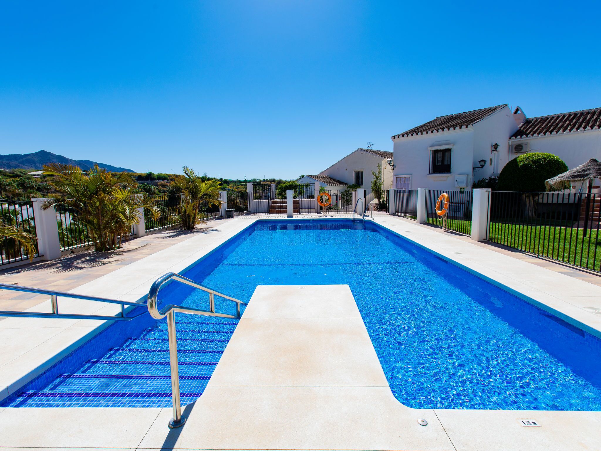 A rectangular swimming pool with a tiled deck, surrounded by a fence, and adjacent to a white building with a tiled roof.