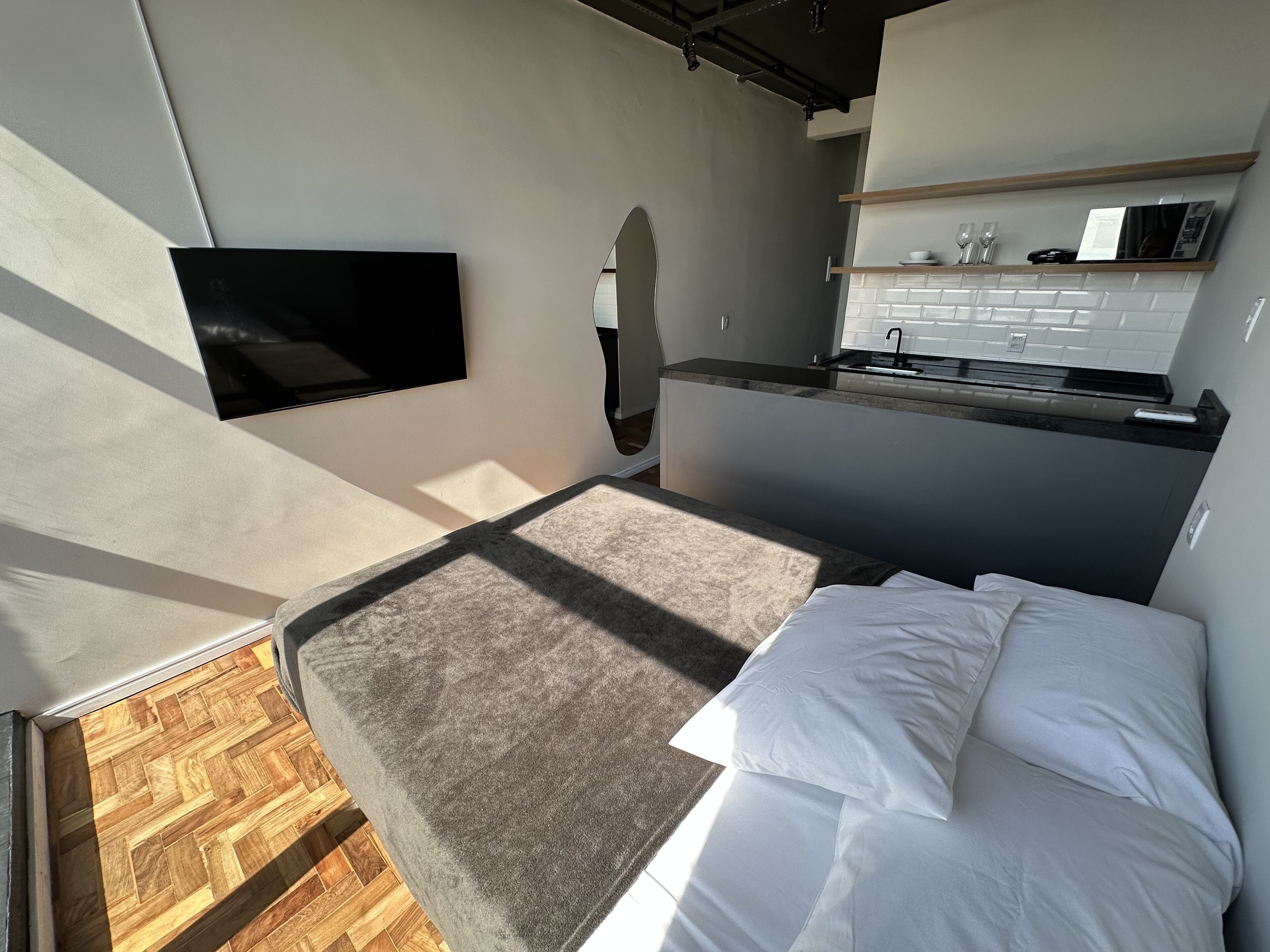 A compact bedroom with a bed, a TV mounted on the wall, and a kitchenette area with a microwave and sink.