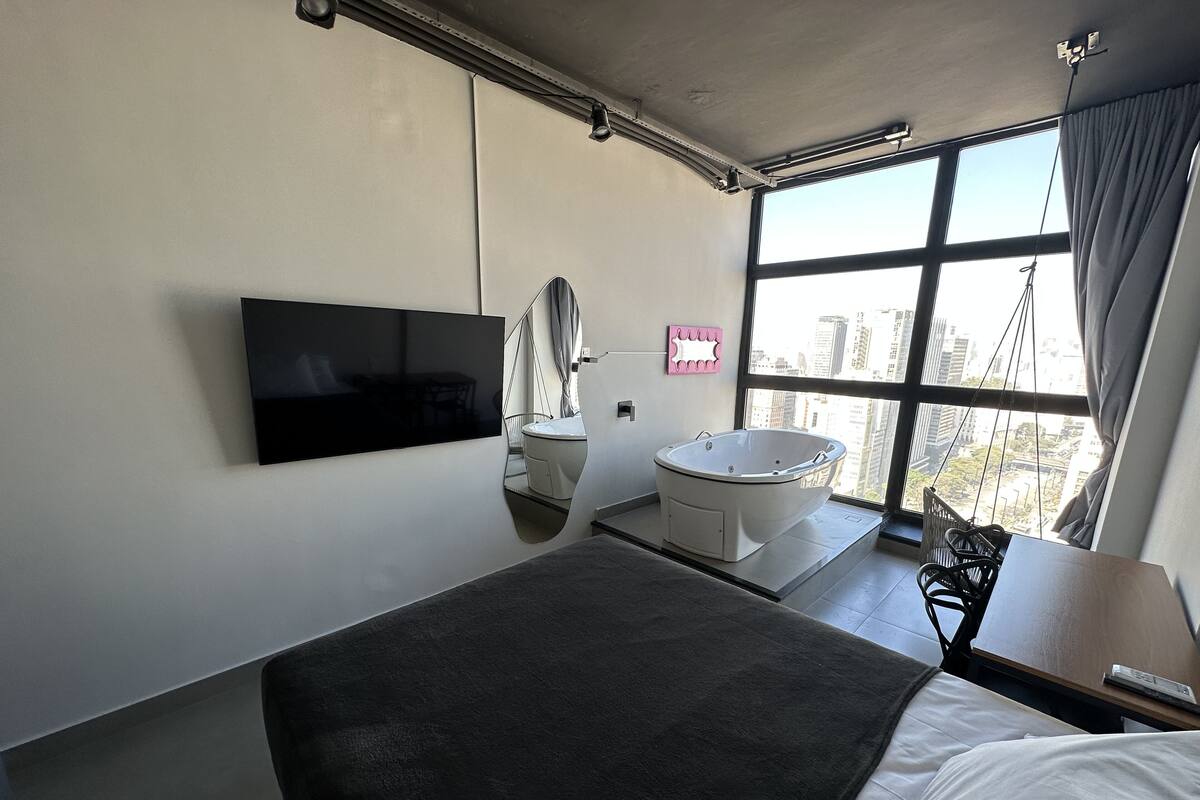 A modern hotel room with a large bed, a mounted TV, a bathtub, and a city view through large windows.