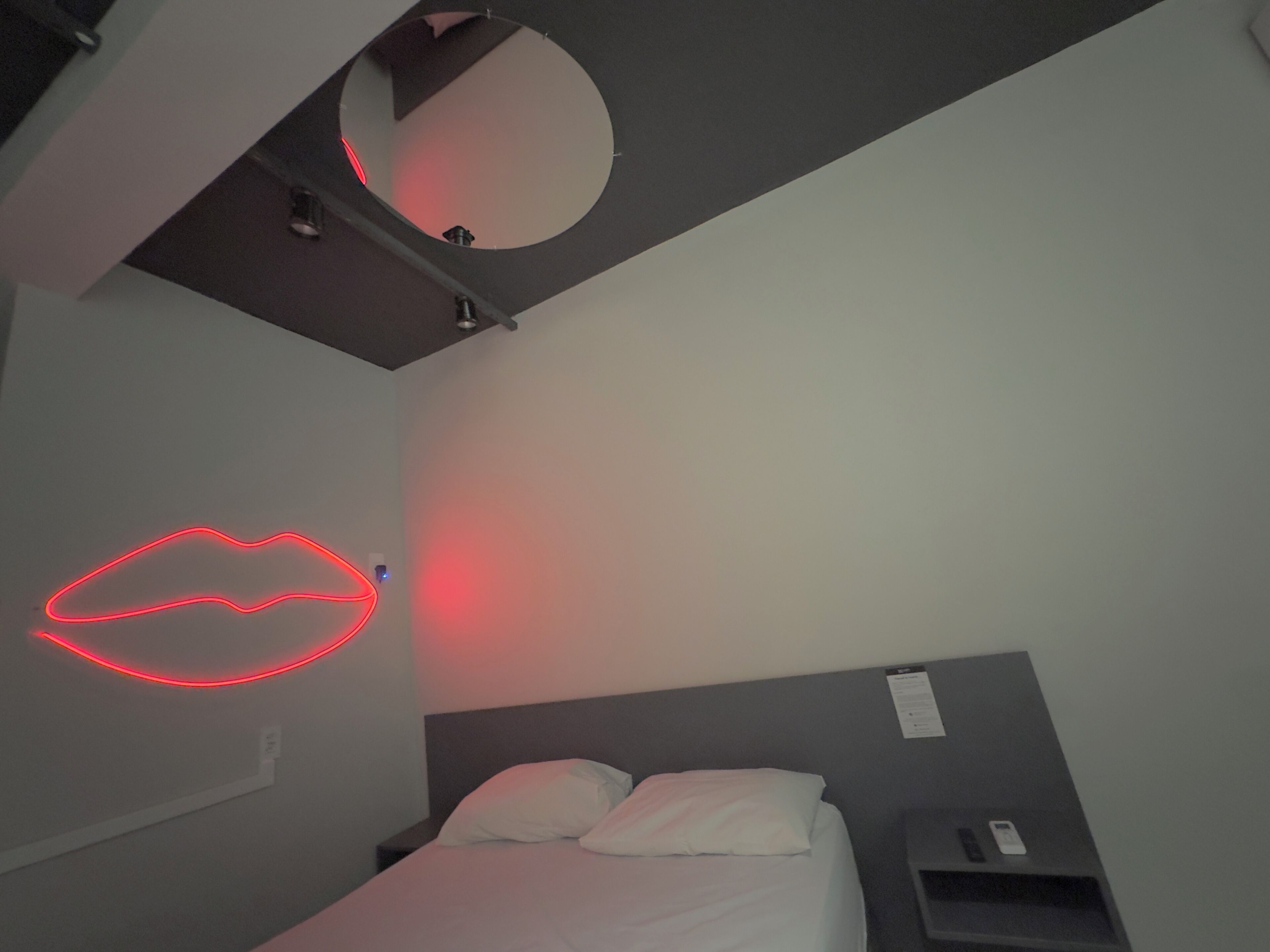 Un dormitorio con cama, mesita de noche, espejo en la pared y una luz de neón roja con forma de labios.