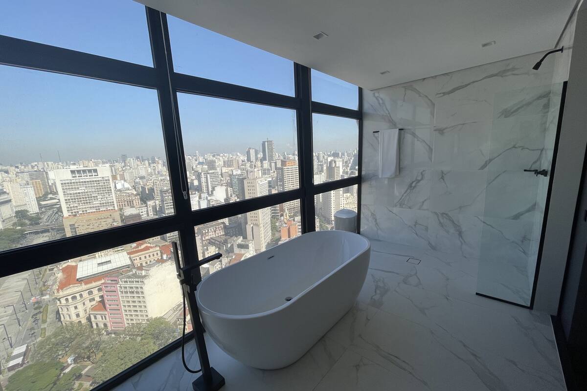 A modern bathroom with a freestanding bathtub, large windows offering a city view, and marble flooring.