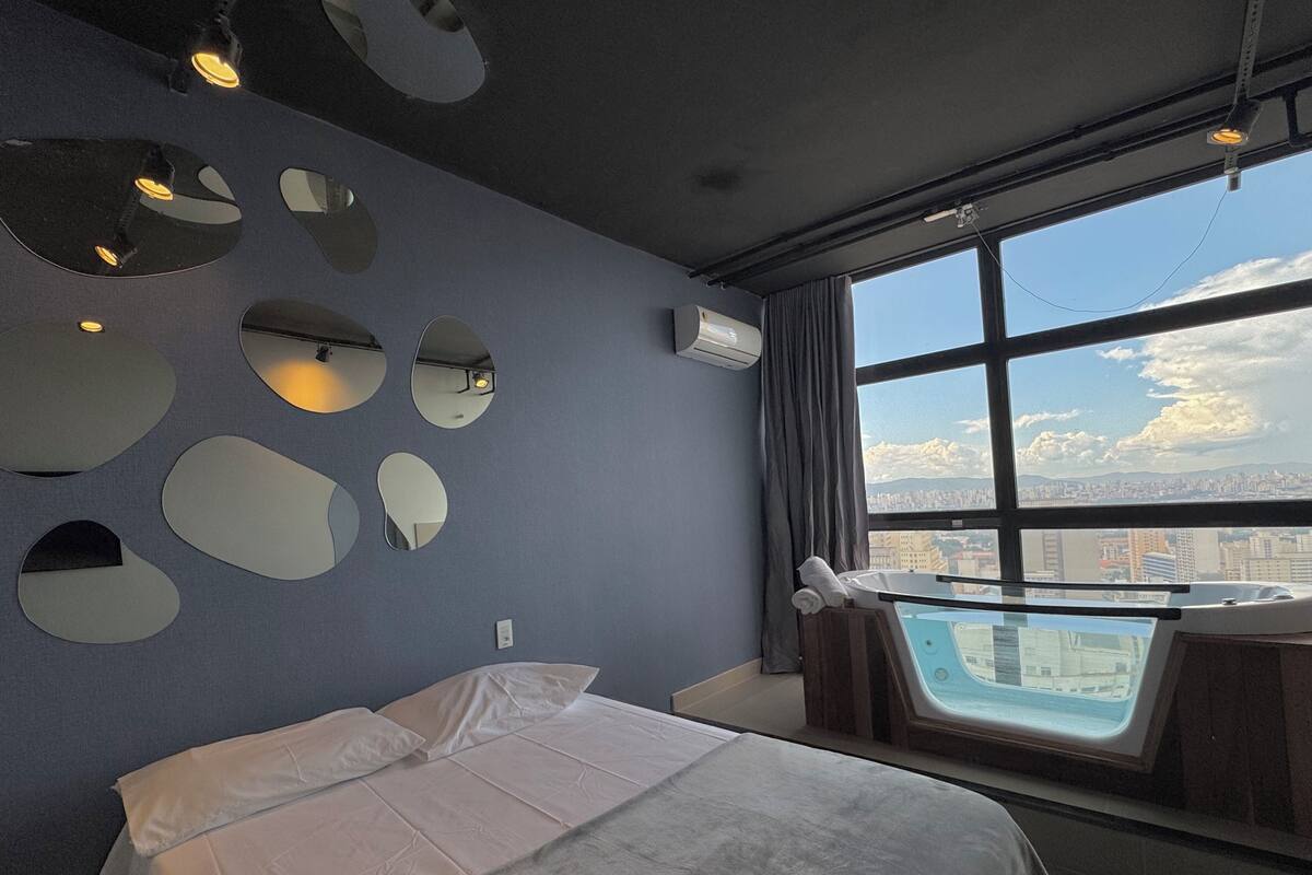 A modern hotel room with a large bed, multiple wall-mounted mirrors, a city view through a large window, and a small seating area with a view.