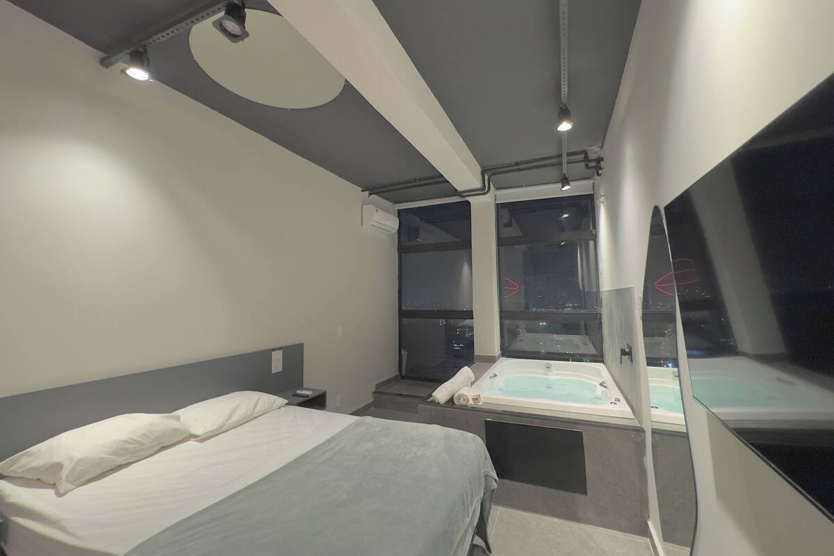 A modern hotel room with a bed, a bathtub, and a large window offering a city view.
