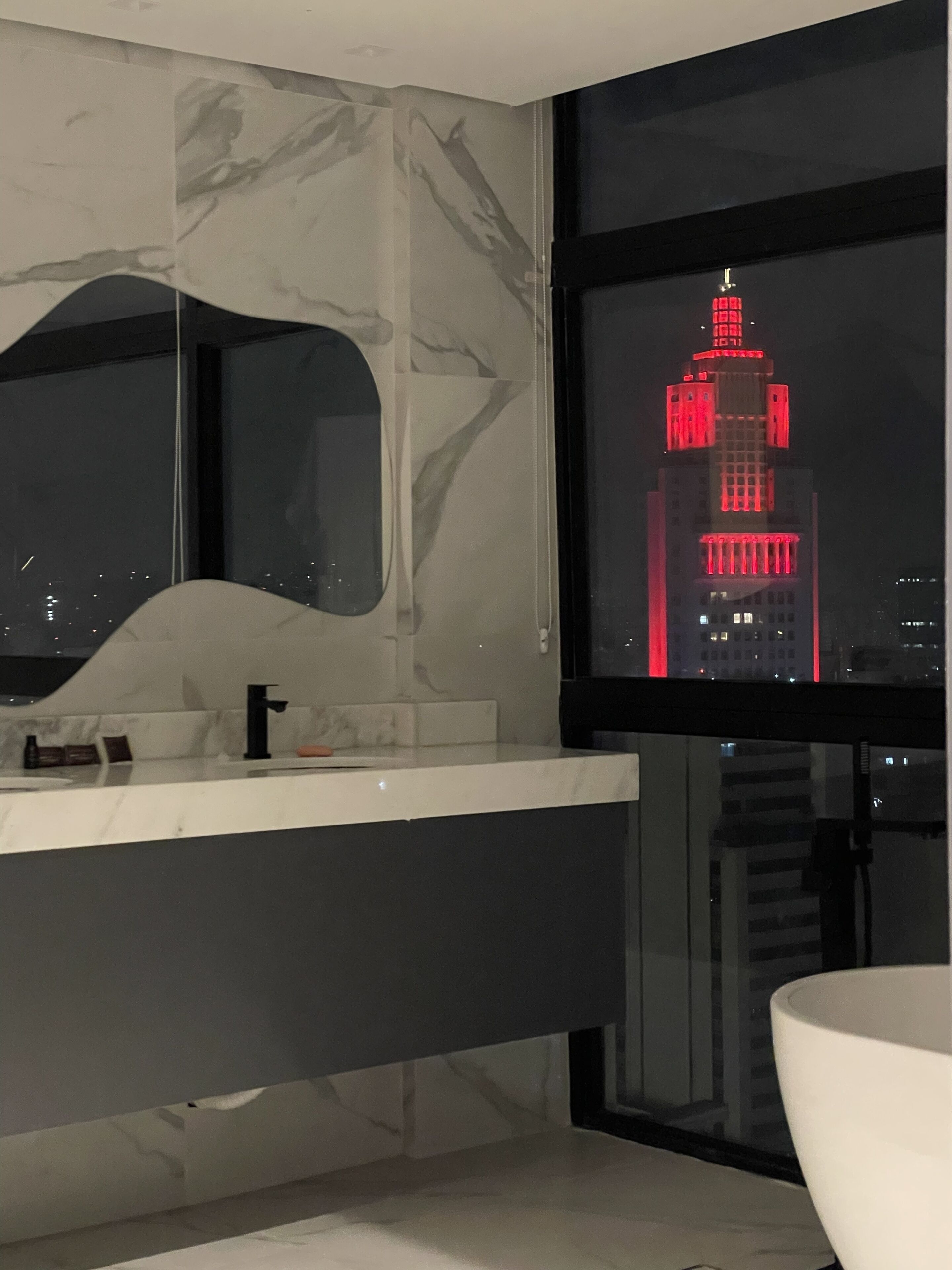 A modern bathroom with a marble vanity, a large mirror, and a window overlooking a cityscape at night.