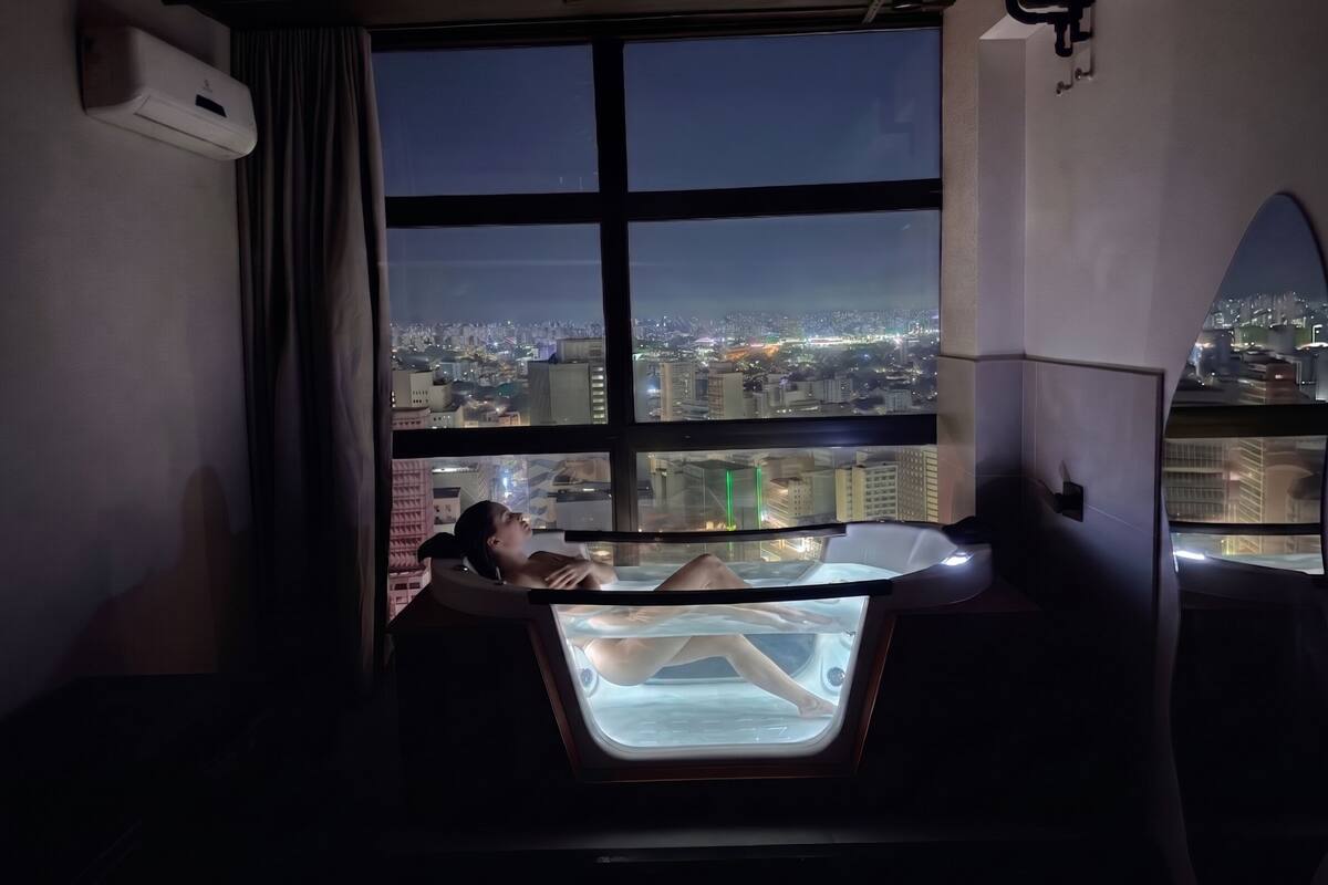 A person lying in a bathtub with a cityscape view through a large window.