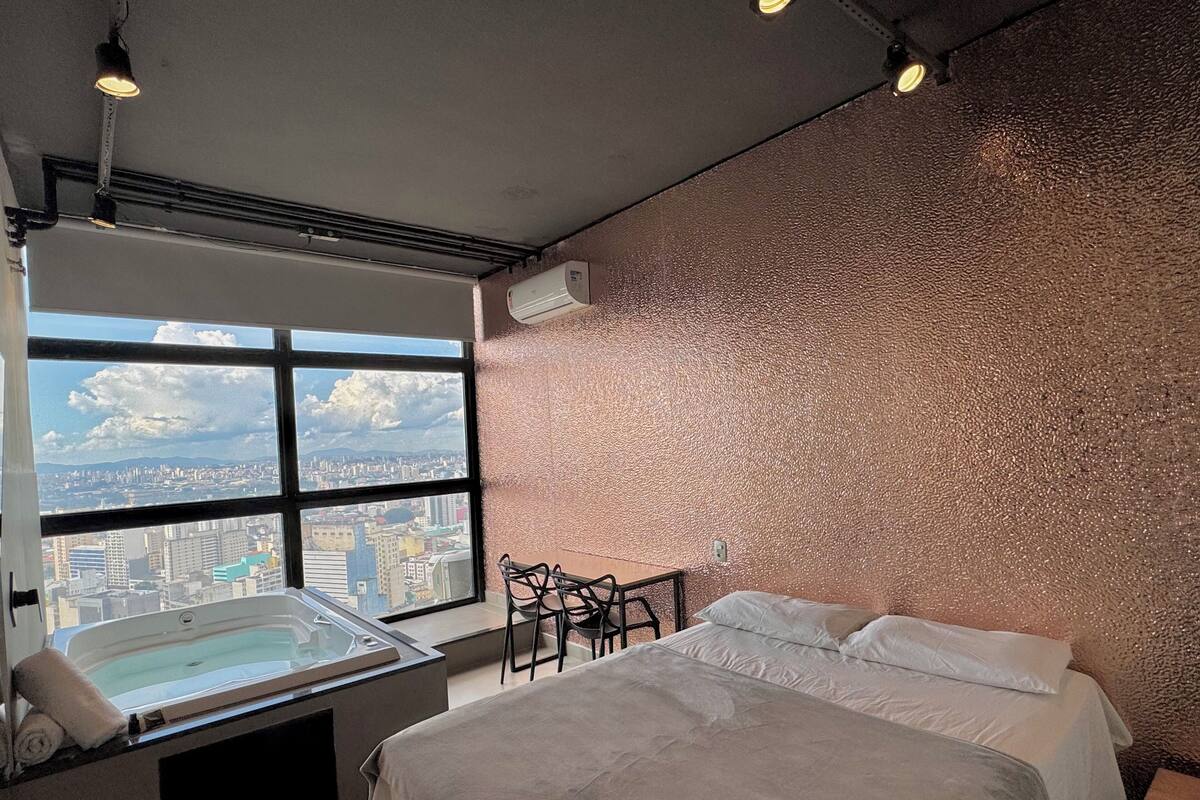 A hotel room with a large window, a bed, a small table, and a chair.