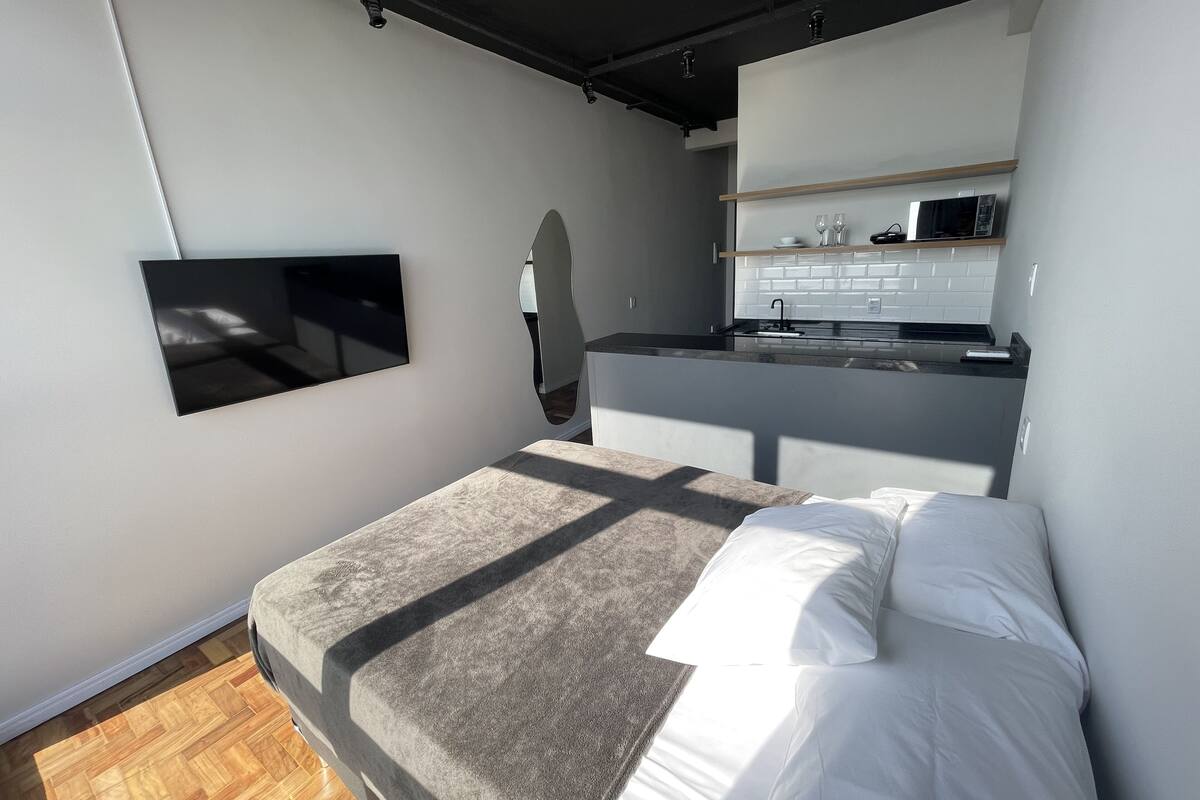 A modern bedroom with a bed, a TV mounted on the wall, and a kitchen area visible through an archway.