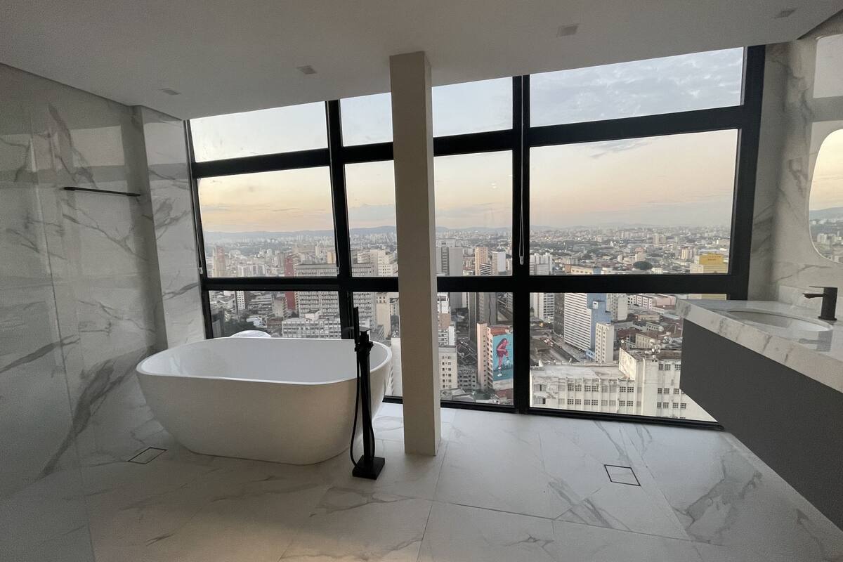 A modern bathroom with a freestanding bathtub, large windows offering a city view, and a marble floor.
