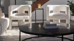 Ein modernes Wohnzimmer mit zwei weißen Sesseln, einem Glastisch und einem kleinen Beistelltisch mit einem dekorativen Gegenstand. Im Hintergrund ist eine Treppe mit Holzwerteln zu sehen.
