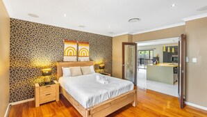 A modern bedroom with a wooden bed, a nightstand, a wall with a giraffe print, and a view of the kitchen through an open door. - Stylish 2BR House for a Memorable Stay @Stretton (Stretton)