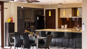 A modern kitchen with dark cabinetry, a central island with bar stools, and a dining area with a red leather sofa. - Stylish 2BR House for a Memorable Stay @Stretton (Stretton)