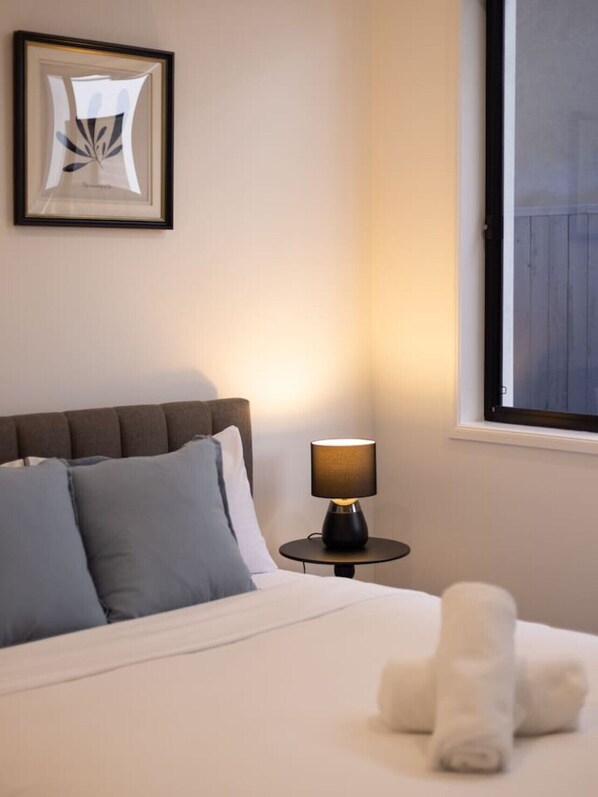 A neatly made bed with a white comforter and blue pillows, a round table with a lamp, a framed abstract artwork on the wall, and a window with a view of a wooden structure.