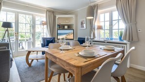 A dining area with a wooden table set for four, a television on the wall, and a view of the outdoors through large windows.
