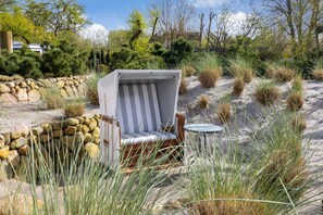 A beachside lounge area with a wicker chair and a small round table, surrounded by sand and sparse vegetation. - Gaat 7 Coquette - Gaat 7 - Wohnen im Historischen Ortskern Keitum (Sylt)