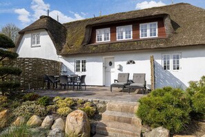 Ein traditionelles Reetdachhaus mit weißer Fassade, einer Steinmauer und einer Terrasse mit Gartenmöbeln.