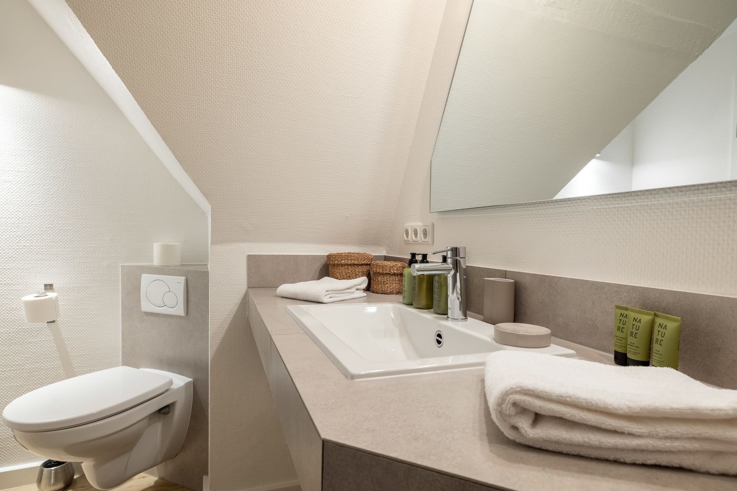 Une salle de bain moderne avec des toilettes blanches, un lavabo surmonté d’un miroir et des articles de toilette posés sur le comptoir.