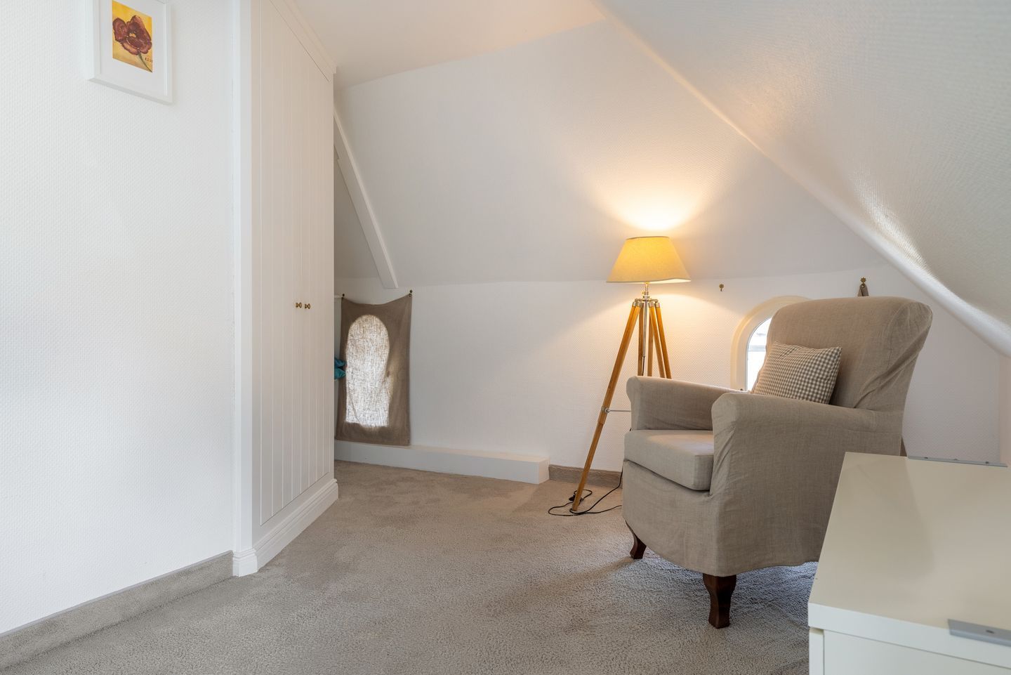 A cozy attic bedroom with a high ceiling, a single window, a floor lamp, a chair, and a small table.