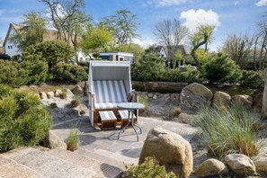 A sunlit outdoor seating area with a striped lounge chair and table, surrounded by rocks and greenery. - Gaat 7 Der Junge Heinrich - Gaat 7 - Wohnen im Historischen Ortskern Keitum (Sylt)