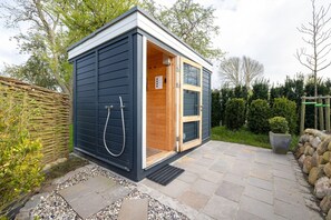 Eine kleine, dunkelblaue Außen-Sauna mit Holztür und an der Wand befestigtem Duschschlauch.
