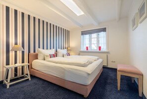 Ein Schlafzimmer mit einem großen Bett, einem Nachttisch, einer Lampe, einem Fenster mit Vorhängen und einer kleinen Bank.