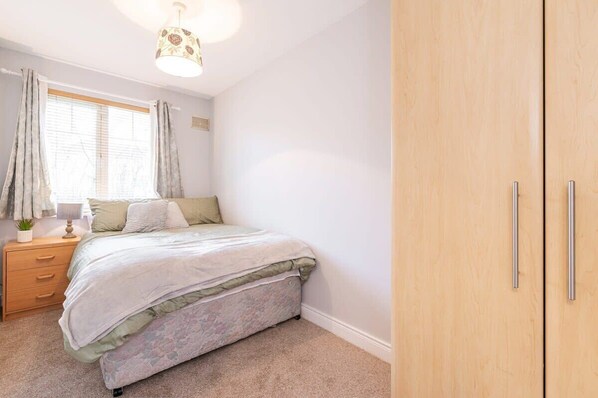 A bedroom with a bed, a nightstand, a lamp, a window with curtains, and a wardrobe.