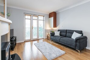 A living room with a grey sofa, a flat-screen TV, a fireplace, and a large window with blinds.