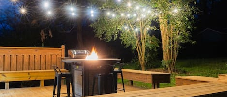 An outdoor deck with a fire pit, string lights, and wooden benches.