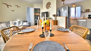 A dining table set with plates, cutlery, and candles, surrounded by wicker chairs in a well-lit room with a kitchen in the background. - Les Pâquerettes by Interhome (Ile d'Oléron)