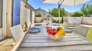 An outdoor patio with a wooden table, blue plates, a fruit basket, and a large umbrella. - Les Pâquerettes by Interhome (Ile d'Oléron)