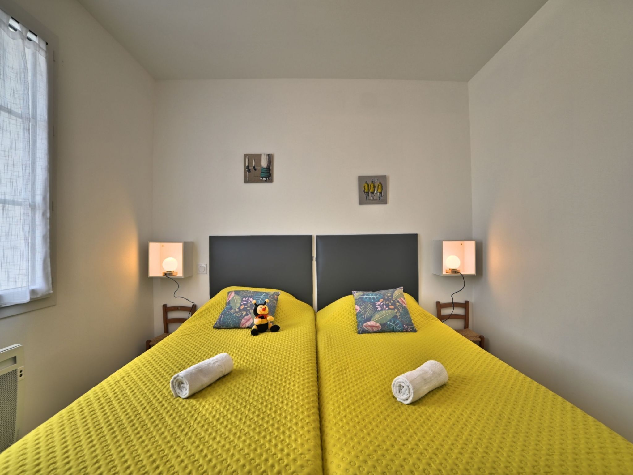 Habitación doble con dos camas, cada una con una colcha amarilla con textura, dos lámparas de pared y un pequeño cuadro sobre cada cama.