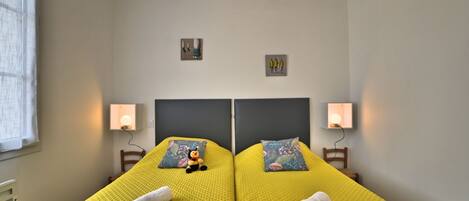 Habitación doble con dos camas, cada una con una colcha amarilla con textura, dos lámparas de pared y un pequeño cuadro sobre cada cama.