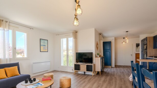 A modern living room with a sofa, coffee table, and TV stand. There is a dining area with blue chairs and a kitchen with blue cabinets.