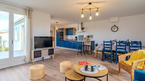 A modern apartment with a living room, dining area, and kitchen. There is a TV, a dining table with chairs, and a kitchen with blue cabinets.