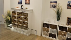 Ein Zimmer mit einem weißen Schrank, der mit 'Manolo's & Slippers' beschriftet ist, inklusive Schubladen für 'Pinsel' und 'Cremes'. An der Wand hängen Gemälde, und auf einem Regal steht ein Topf mit einer Pflanze.