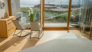 Ein modernes Zimmer mit großem Fenster, zwei Schaukelstühlen, einer Holzkommode und einem Bett mit gesteppter Bettdecke.