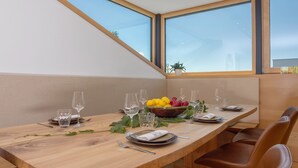Een eettafel gedekt met borden, glazen en een schaal fruit, met uitzicht op de lucht door grote ramen.