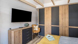 Ein Schlafzimmer mit an der Wand montiertem Fernseher, einem Holzkasten, einem Stuhl und einem Bett mit gelber Decke.