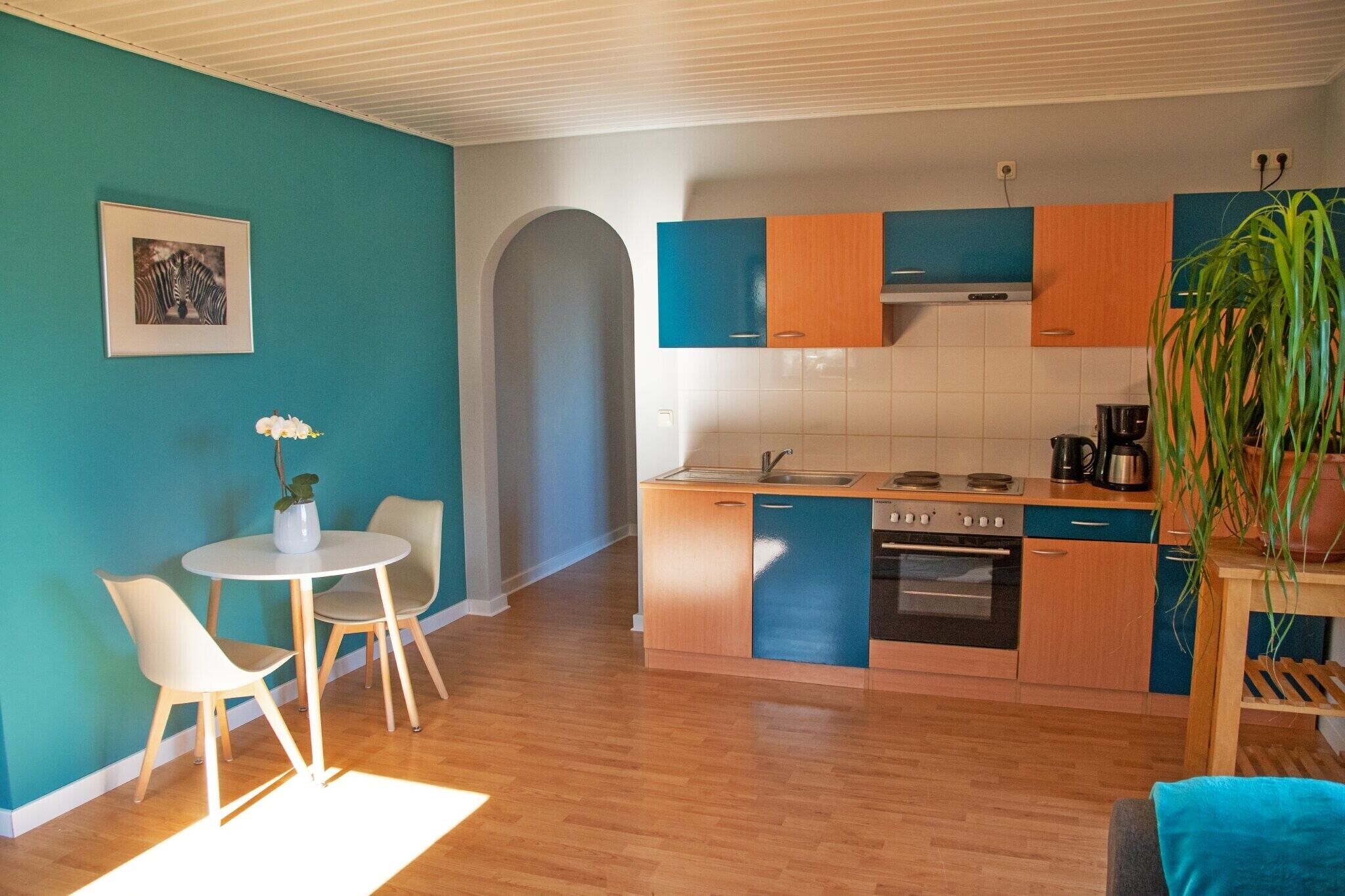 A compact kitchen with a sink, stove, and cabinets, a dining area with chairs, and a potted plant.