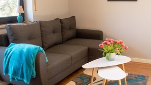 A modern living room with a sofa, a small table with a vase of flowers, a painting on the wall, and a window with a view of trees. - Apartment 'Ferienwohnung Maintalblick' with Private Terrace, Private Garden and Wi-Fi (Eltmann)