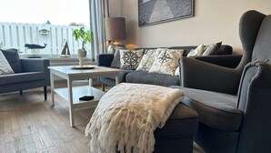 Una sala de estar moderna con un sofá gris, una mesa de centro blanca y un sillón gris. Hay un ventanal con vistas a una valla blanca y un árbol.