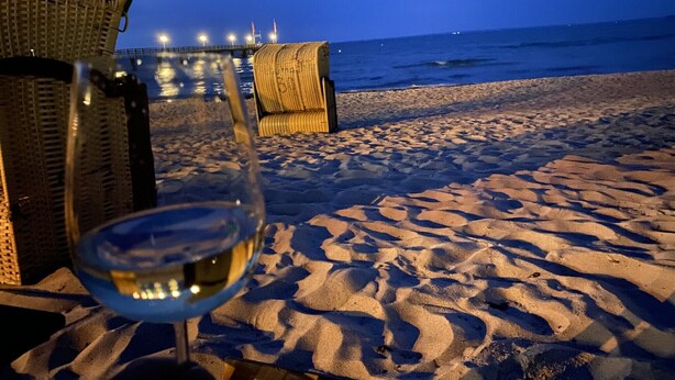 Escena de playa con una silla de mimbre y una copa de vino blanco.