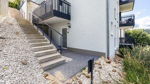 Ein modernes Gebäude mit einer Treppe, die zu einem Balkon führt, einer gepflasterten Fläche und Blick ins Grüne.